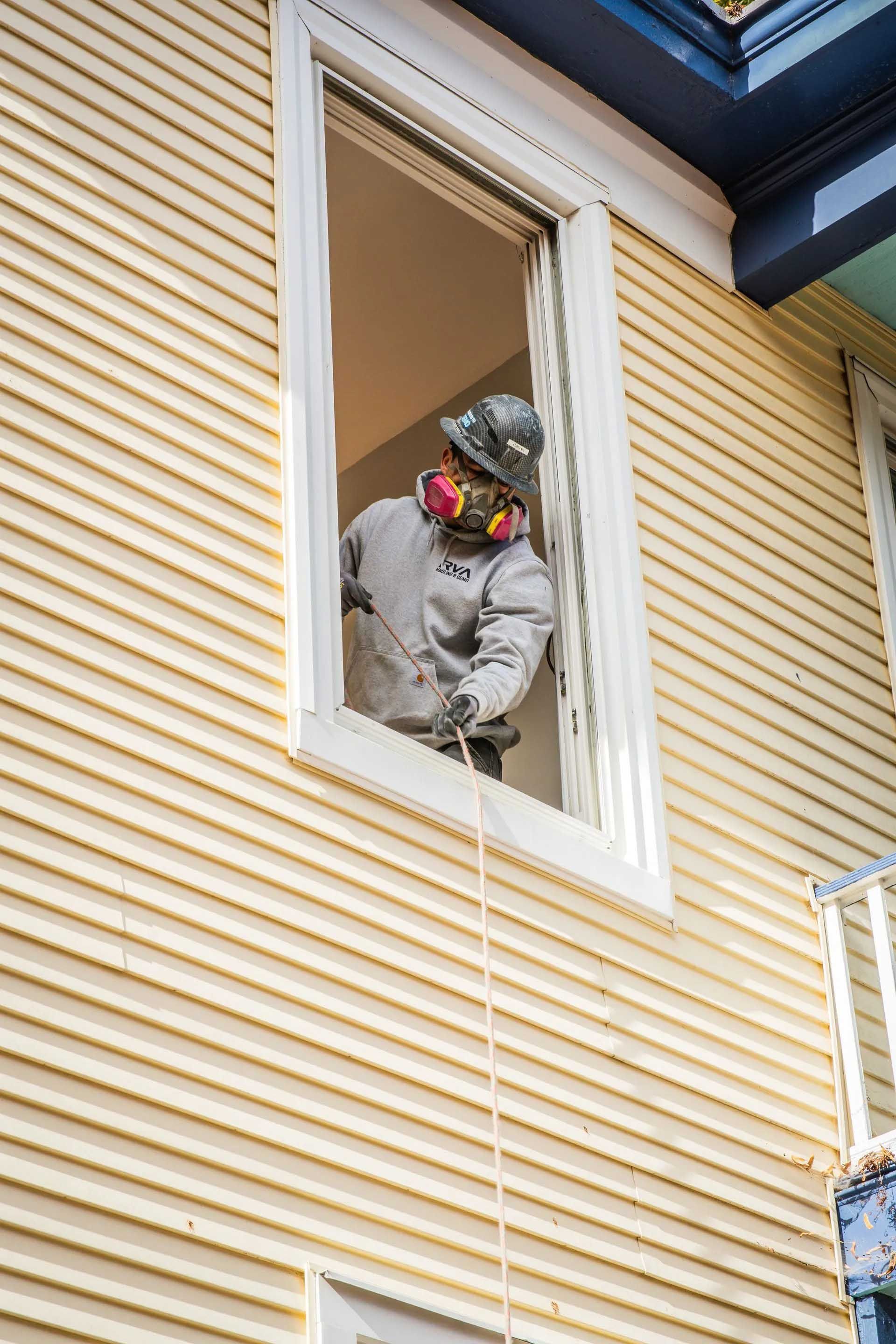 Worker in protective gear leaning out of a second-story window on a yellow house