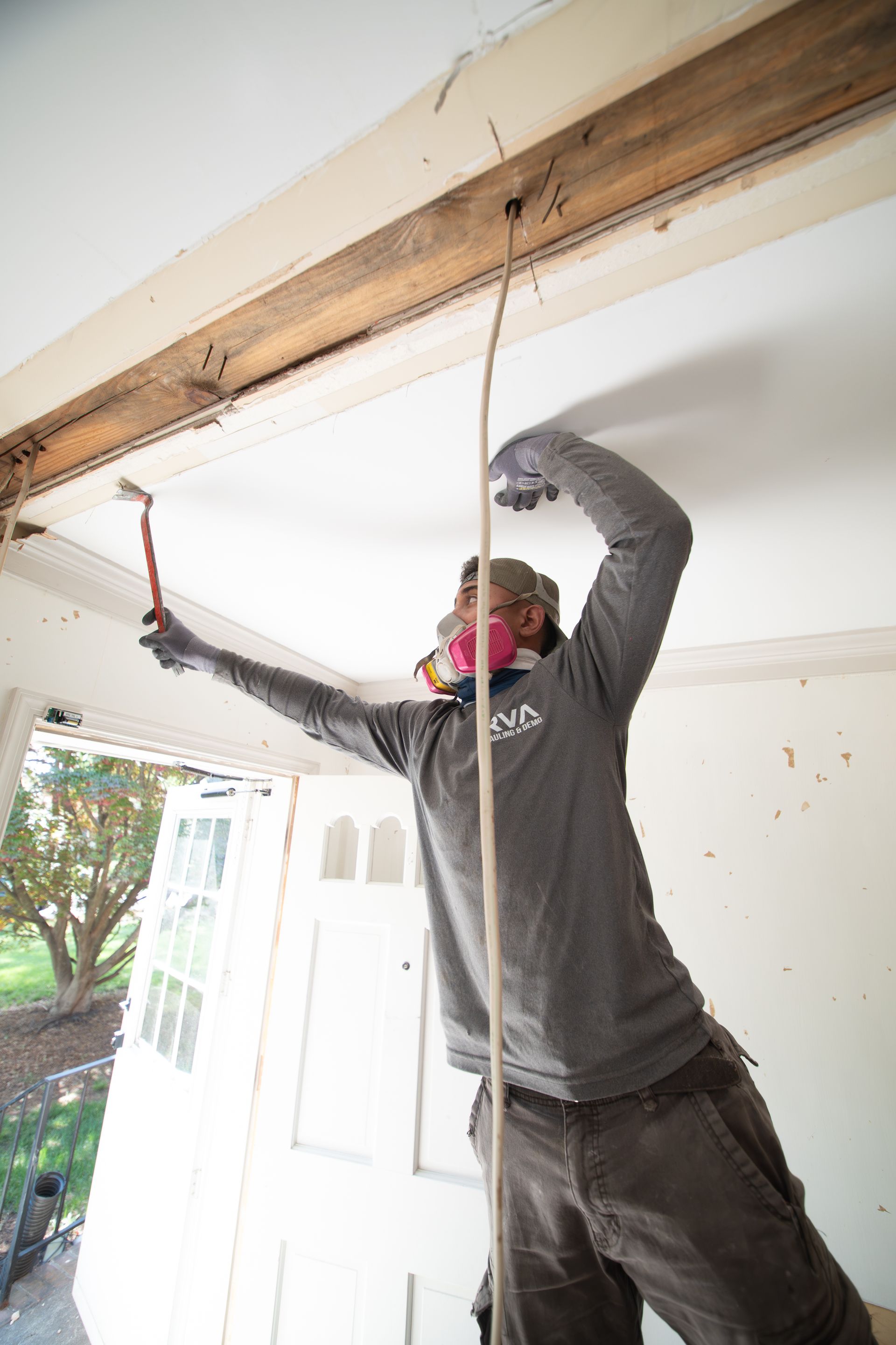 Person in a dark shirt and pants working on exposed ceiling beams with a tool in a room near an open door.