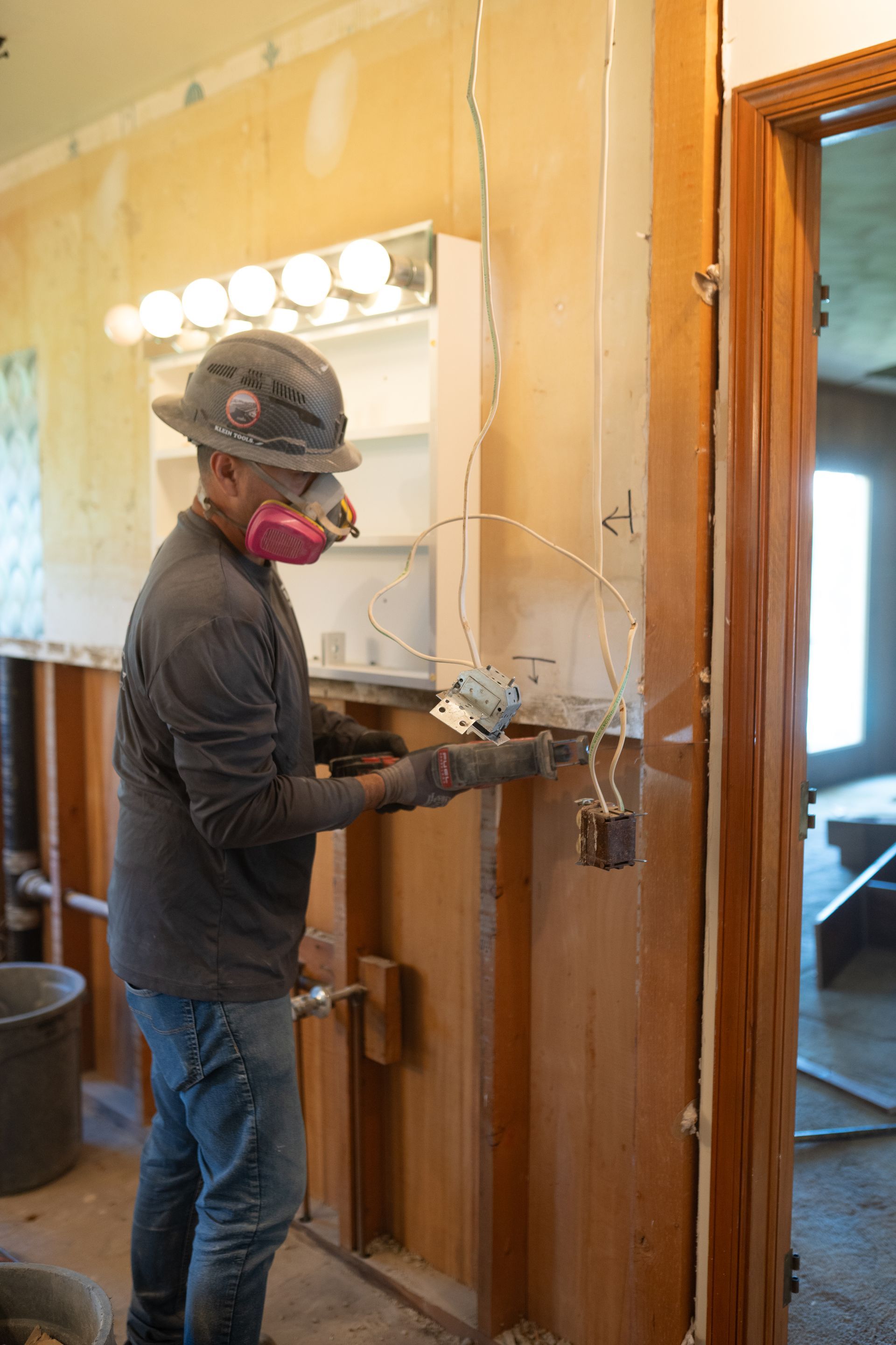 Worker in hard hat and mask drilling into a wall beside a doorway in a room under repair