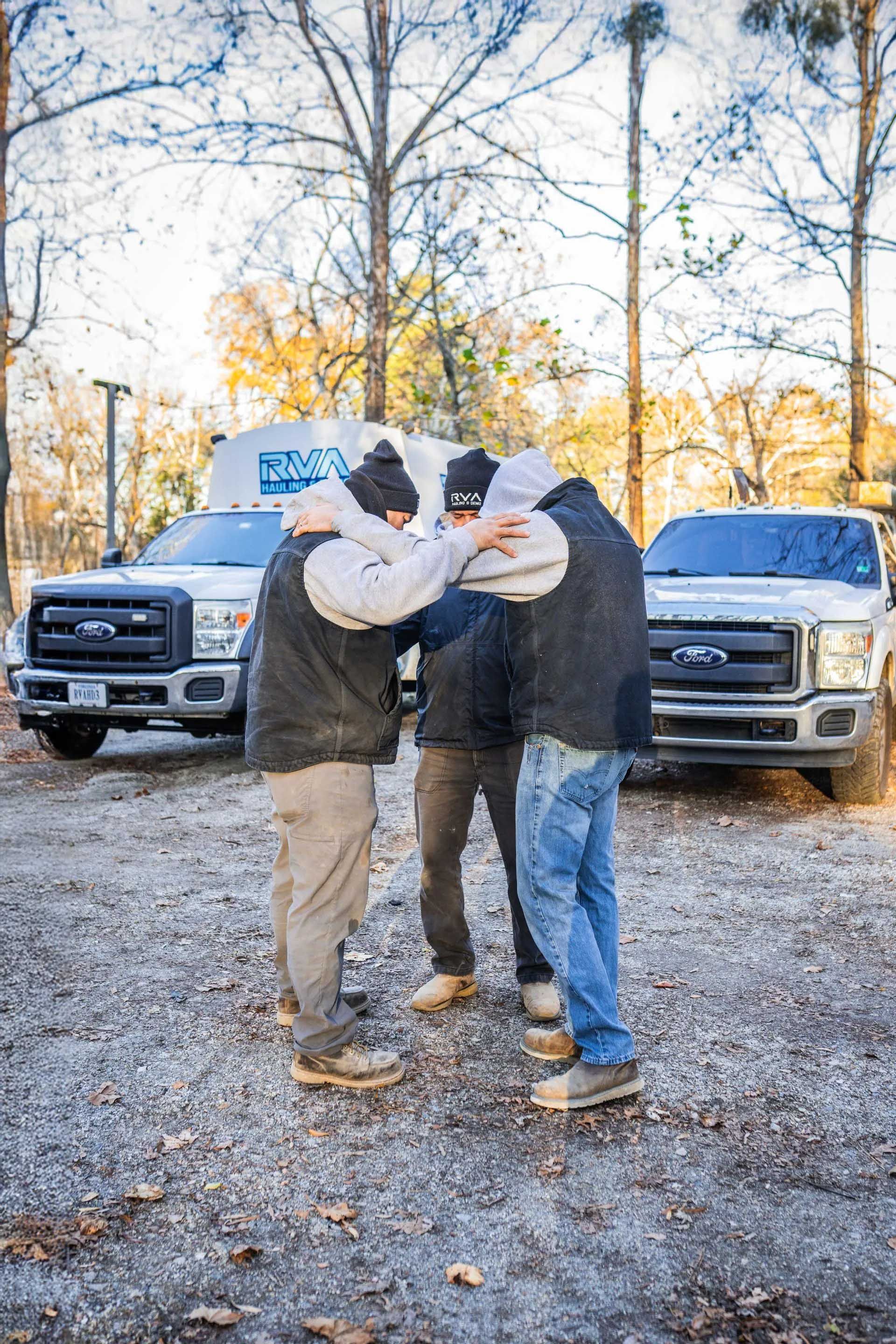 Two people in winter hats embracing in a gravel lot, with pickup trucks and bare trees behind them.