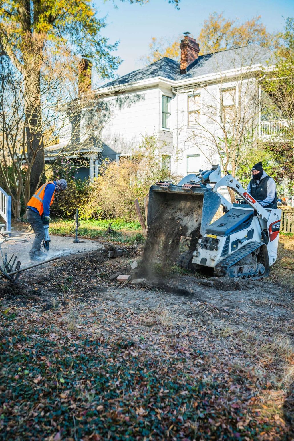 Workers use a skid steer to clear leaves and debris in a yard beside a house