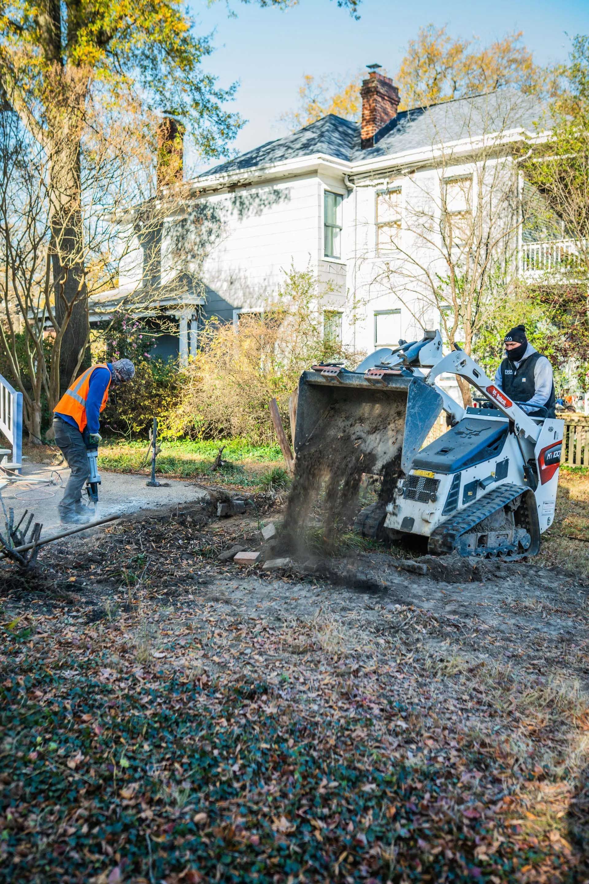 Workers use a skid steer to clear leaves and debris in a yard beside a house