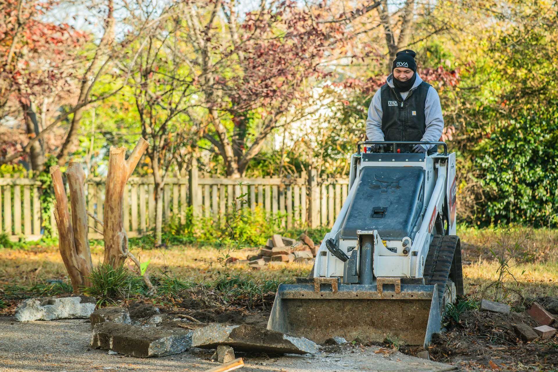 Worker operating a skid steer in a leafy yard, clearing soil near a fence in autumn