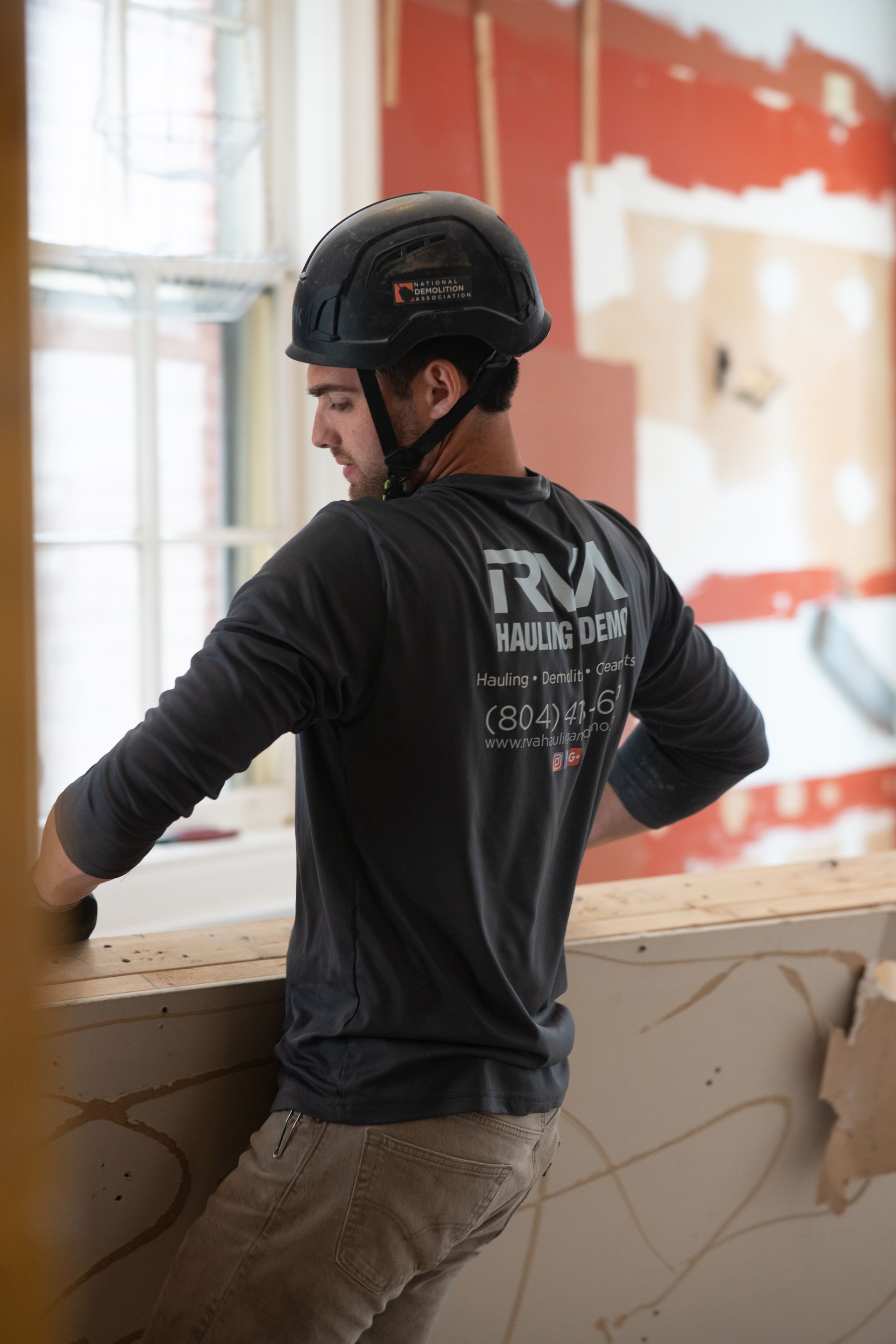 Worker in black shirt and helmet stands by a window in a room under renovation.