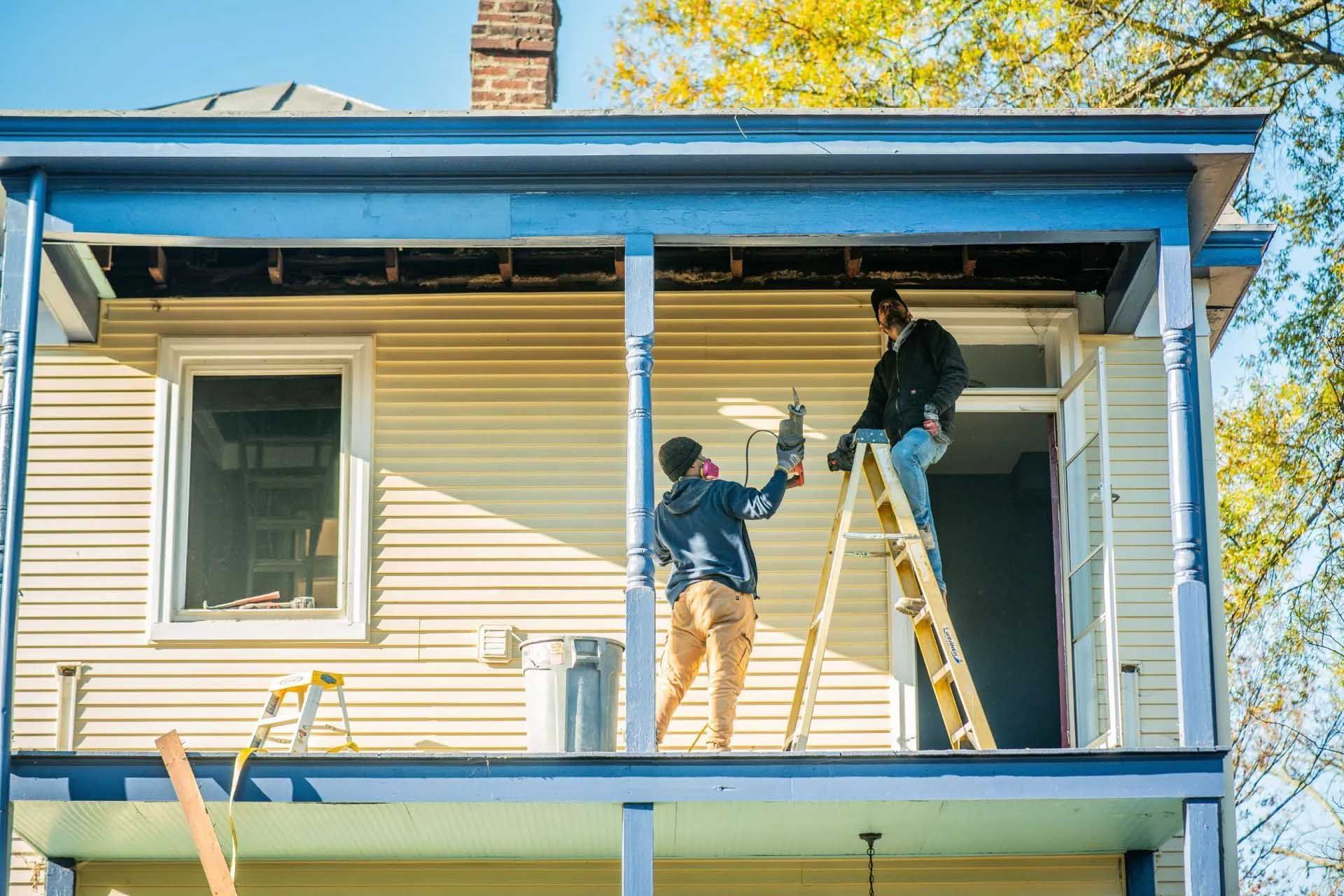 Two workers painting a beige house exterior on a porch ladder under a blue sky