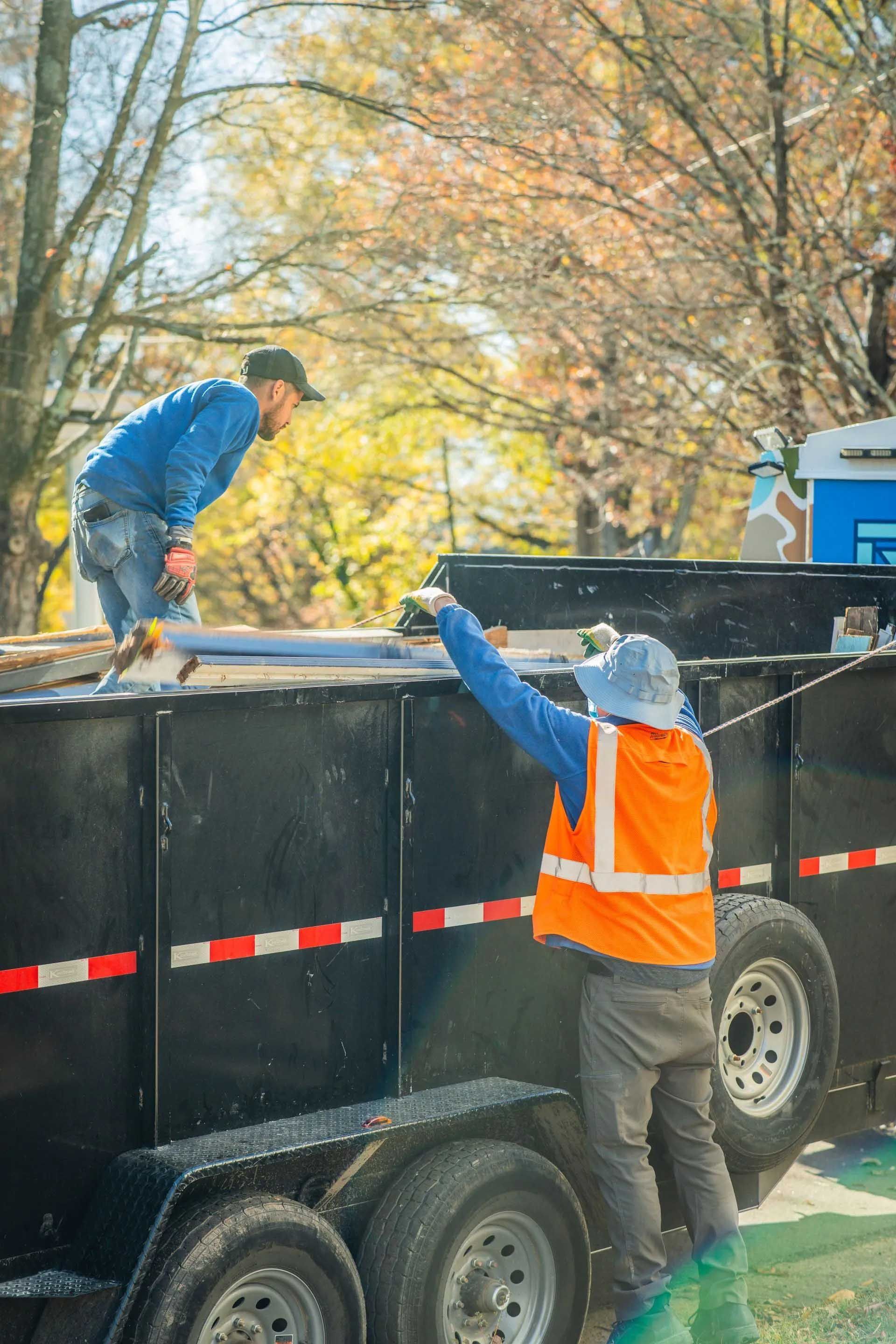 Two workers loading debris into a black dump trailer outdoors, one in a blue jacket and one in an orange vest.