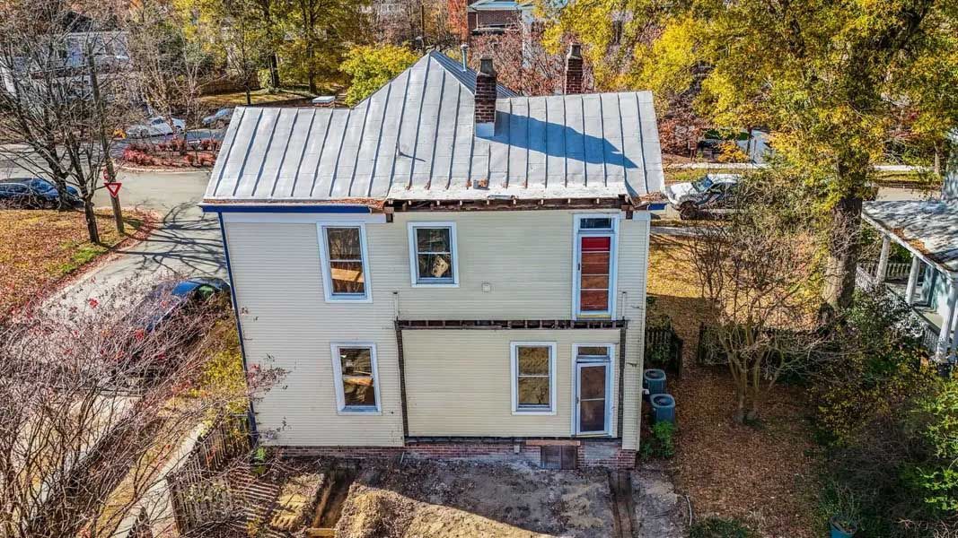 A small white two-story house with a metal roof and surrounding trees, seen from above.