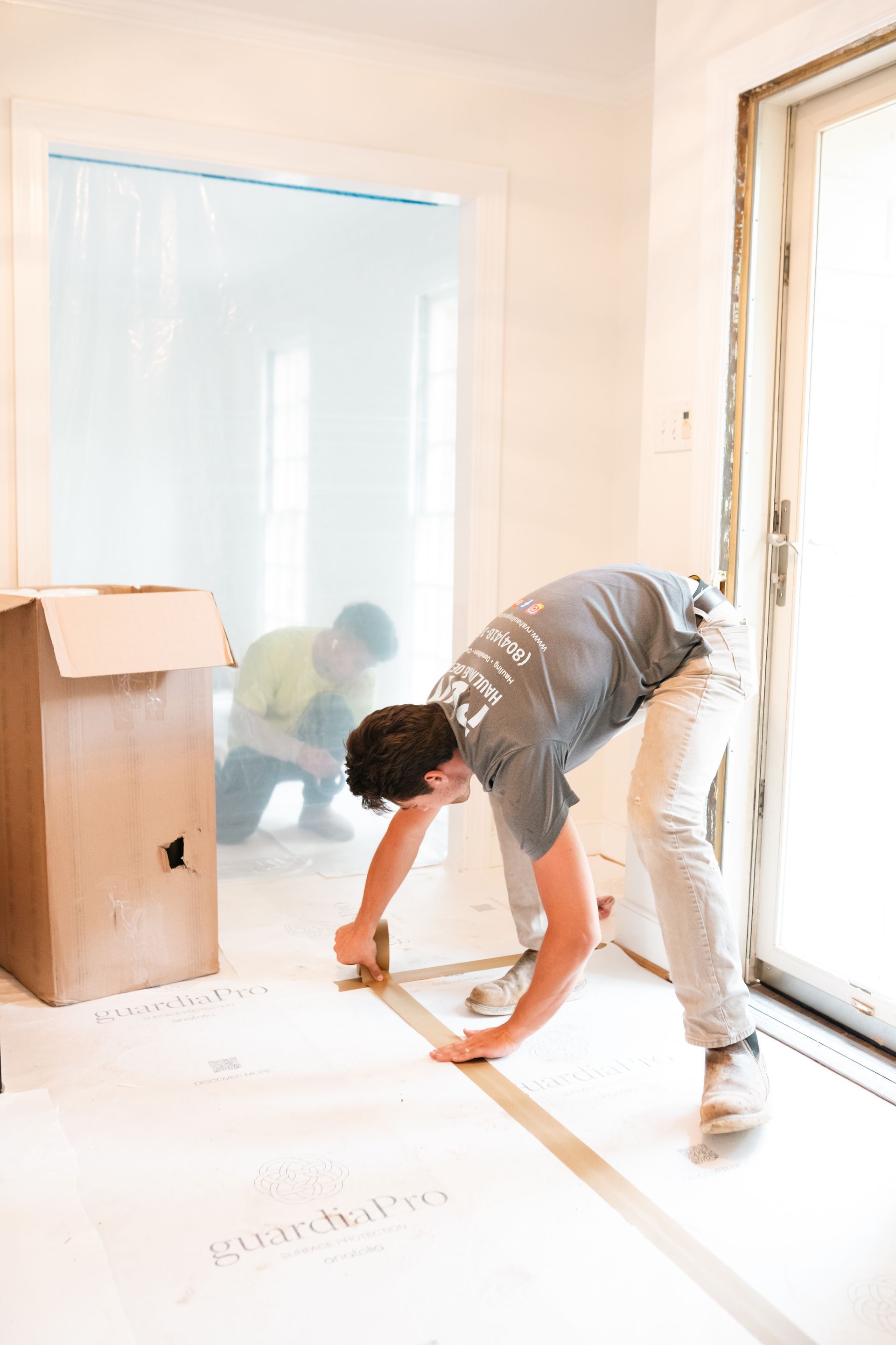 Person bending over in a bright room, reaching toward the floor near a doorway and cardboard box.