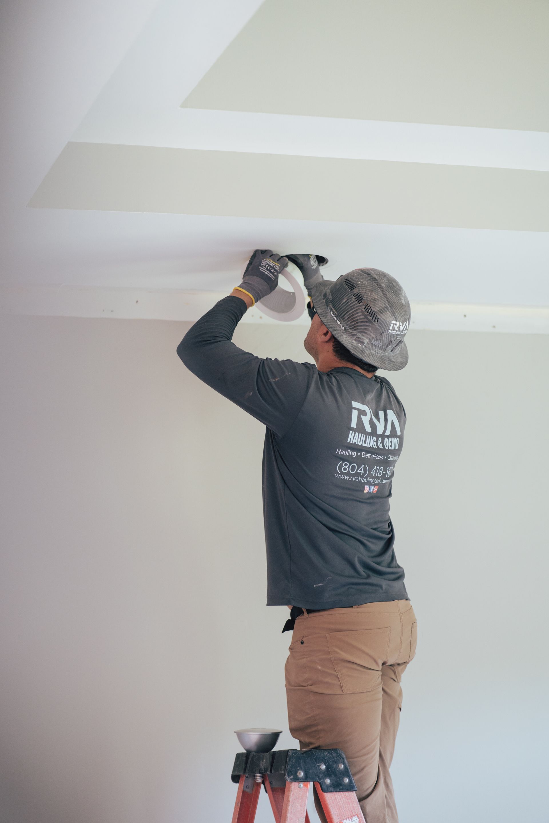 Worker in hard hat painting a white ceiling on a step stool inside a room.