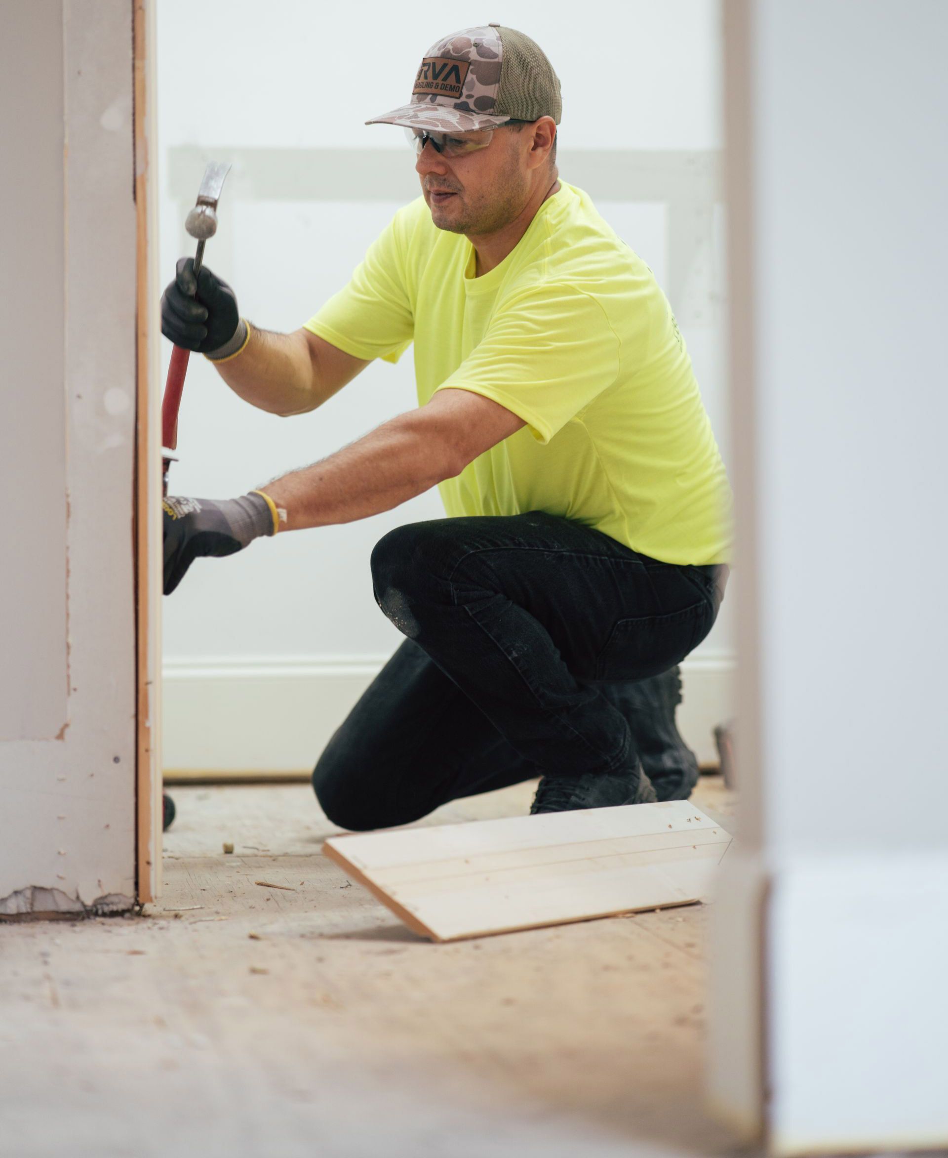 Worker in neon shirt kneeling, using a hammer to install trim in a doorway during home renovation.