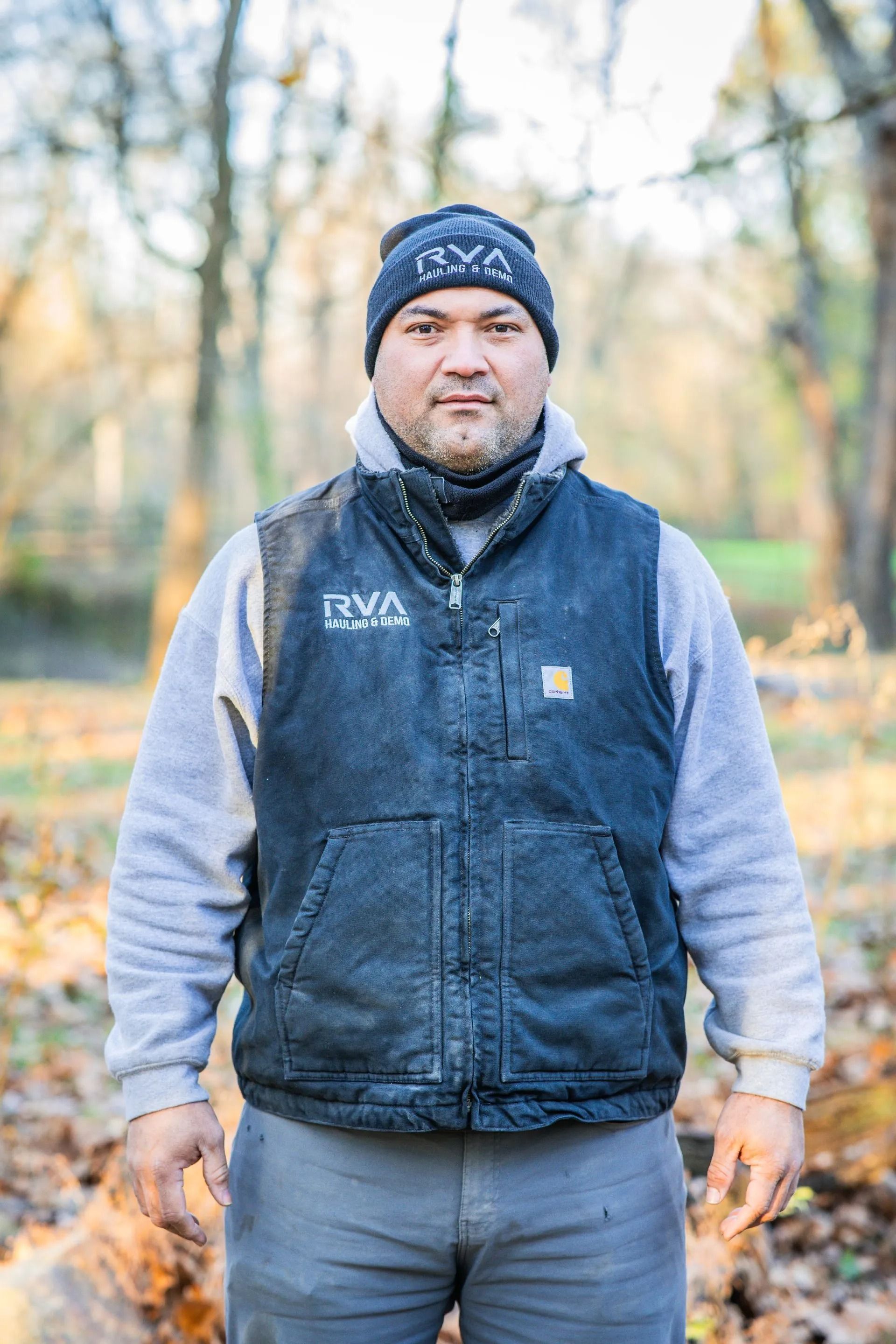Man in a blue vest and knit cap standing outdoors in a wooded park during autumn