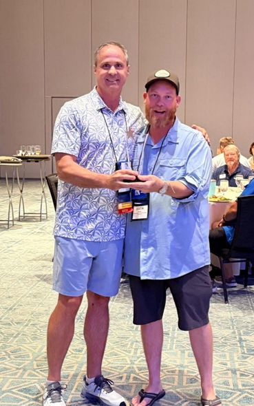 Two men in blue shirts stand indoors, smiling and holding an award plaque at a conference.
