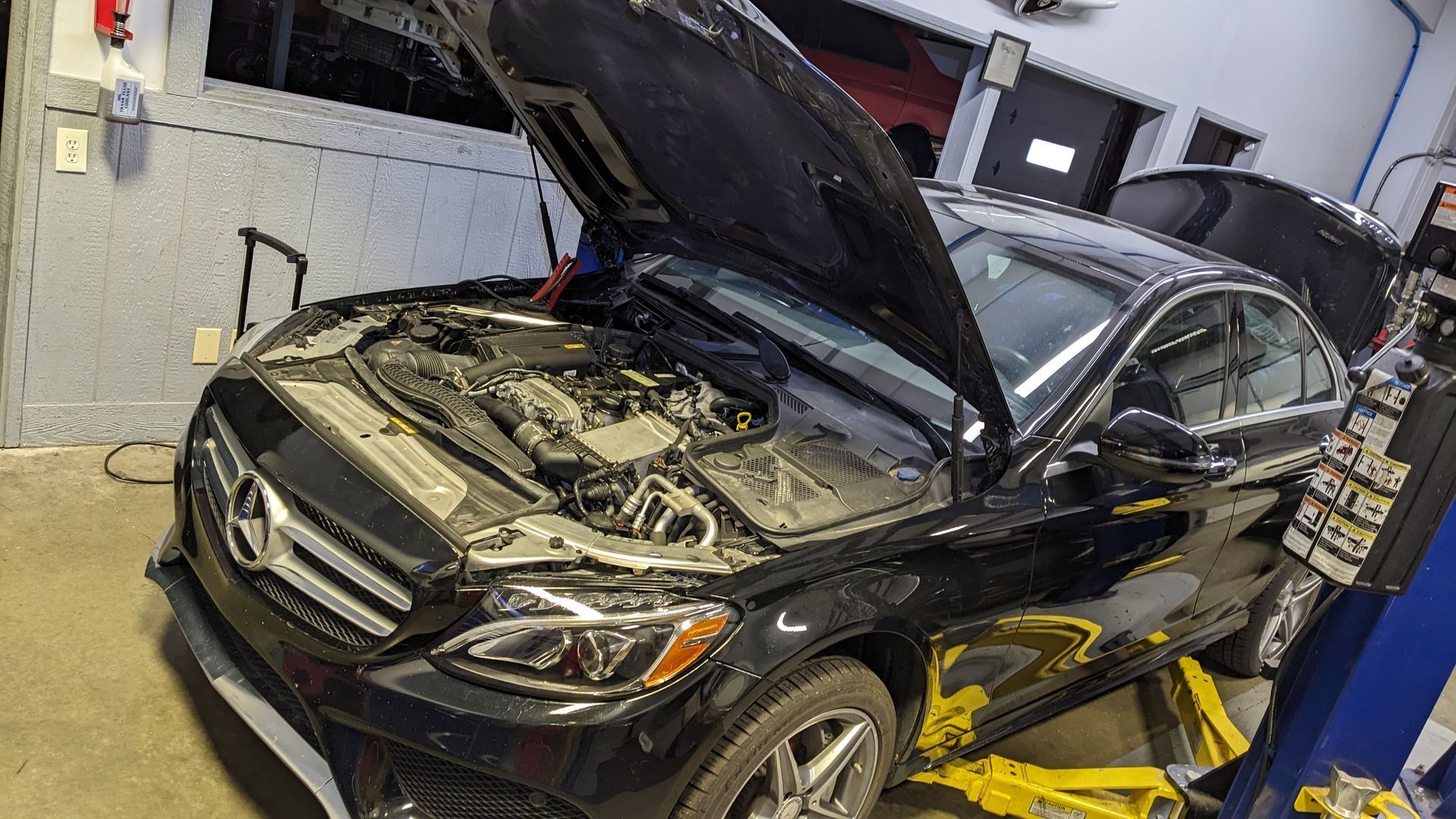 A black mercedes benz with the hood up is on a lift in a garage.