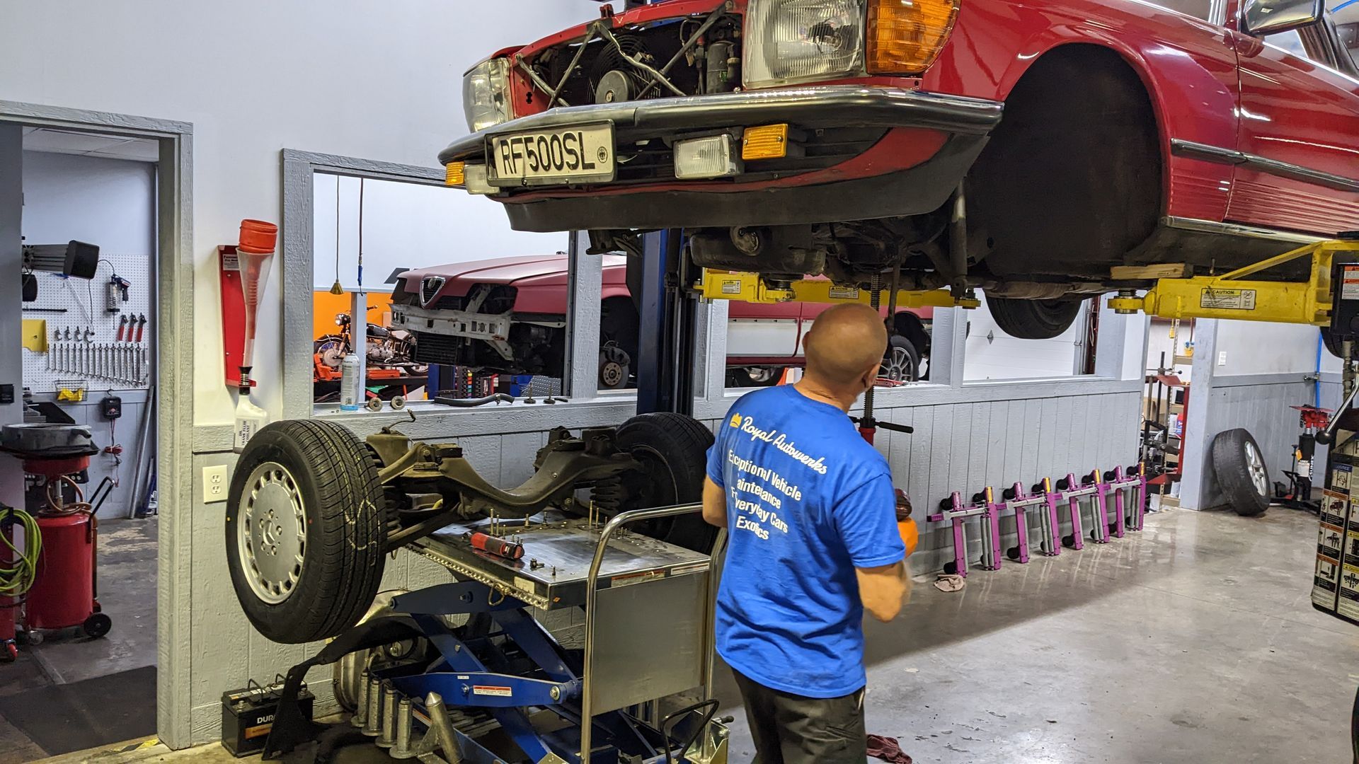 A man in a blue shirt is working on a red car in a garage.