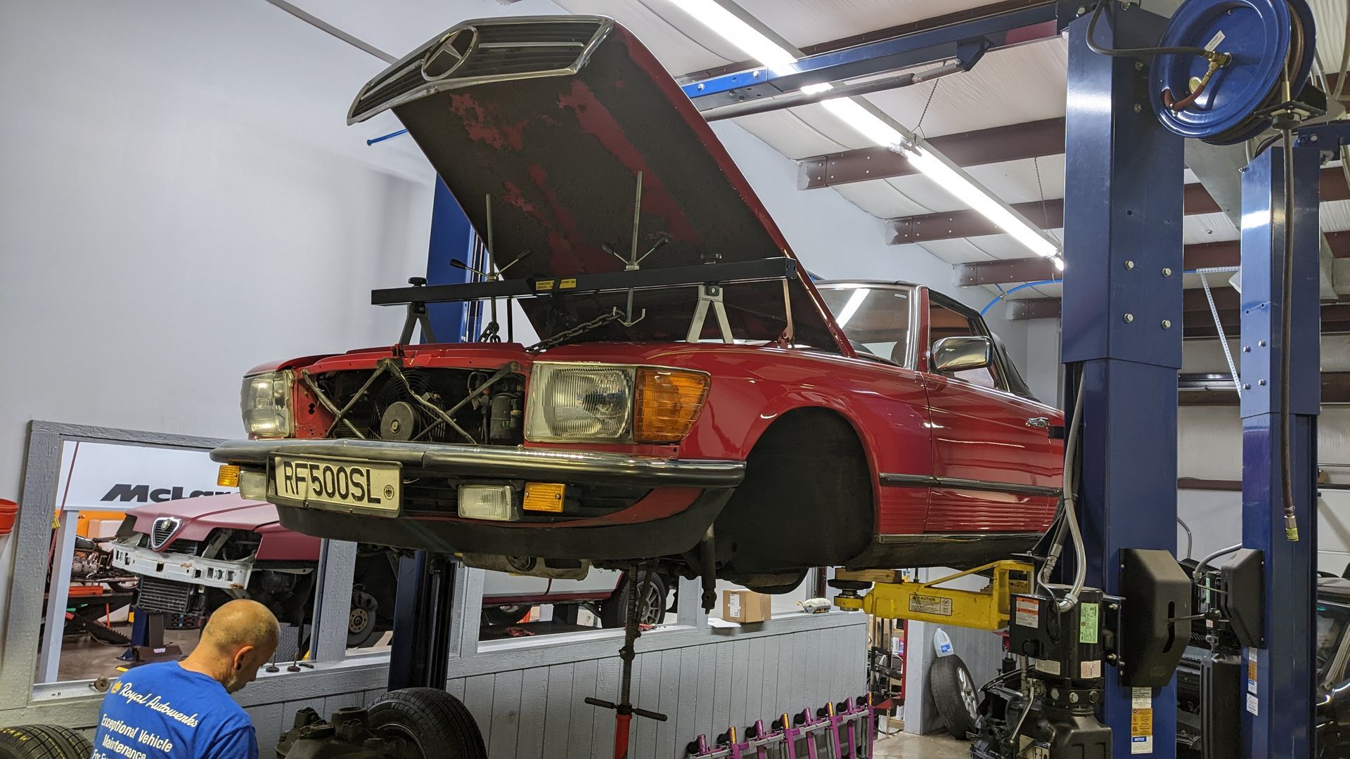 A red car with the hood up is on a lift in a garage.
