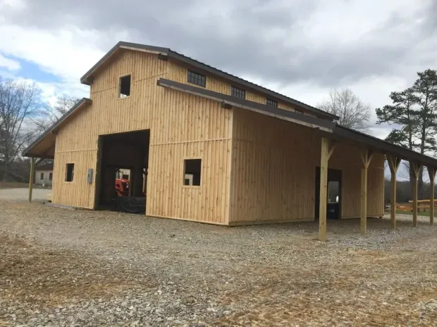 Barn with light-colored wooden siding, open doors, and covered porch on a gravel lot under a cloudy sky.