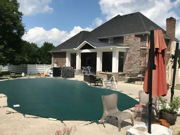 Backyard with pool covered by green tarp, house with brick and stone, patio furniture, and blue sky.