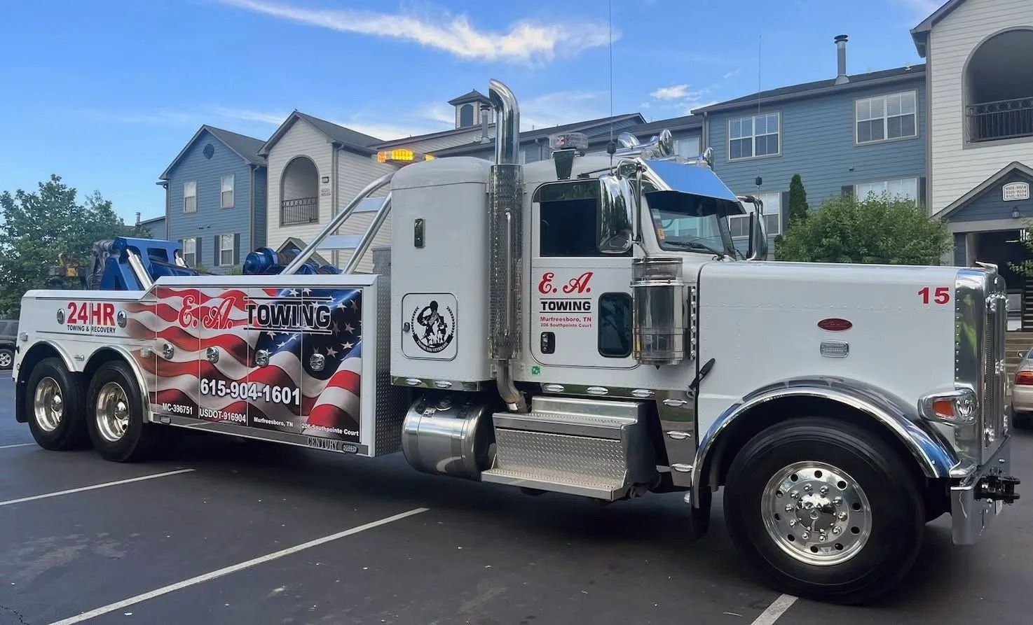 White tow truck with American flag graphics parked in front of apartments on a sunny day.