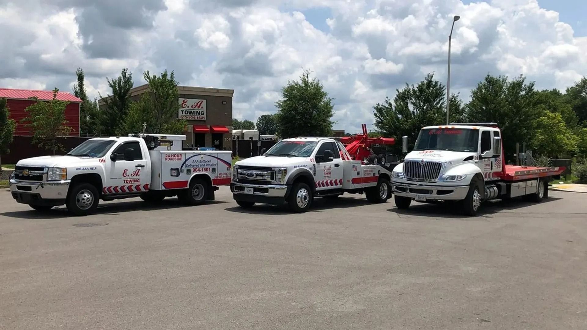 Three white tow trucks parked on asphalt in front of a building on a sunny day.