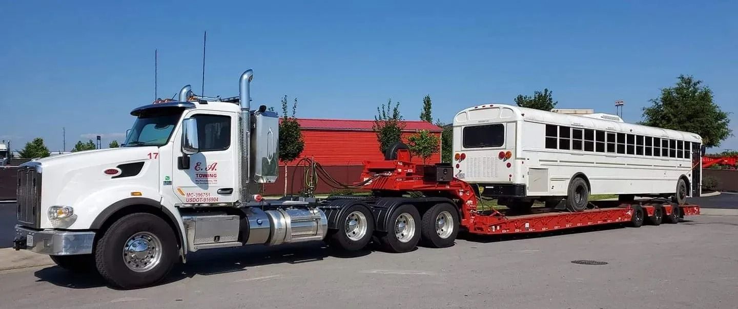 White truck with a bus on a flatbed trailer, set against a blue sky.