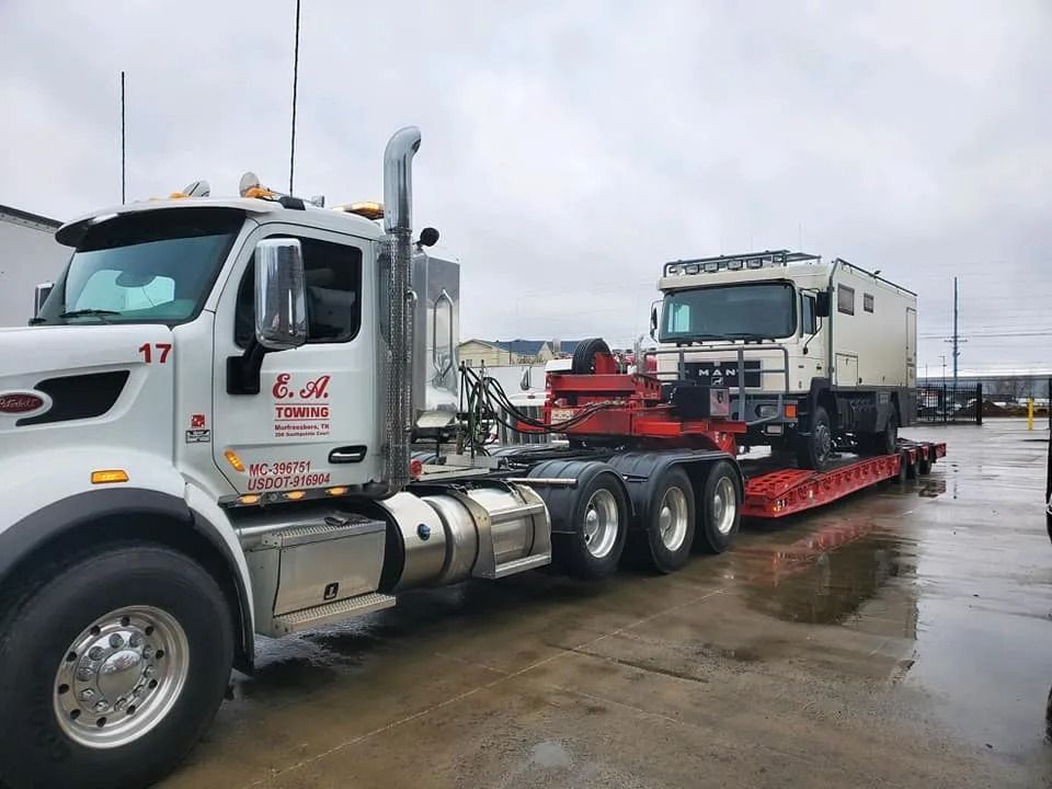 White semi-truck hauling a large light-colored vehicle on a red flatbed trailer, outdoors on a cloudy day.
