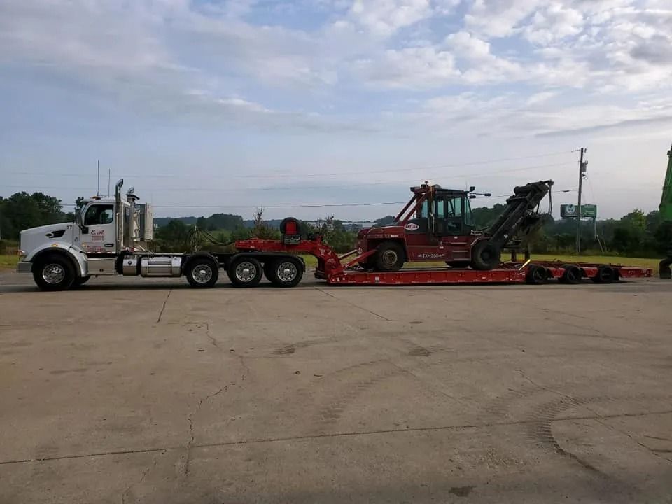 A white semi-truck with a red trailer carrying a red forklift on a paved lot under a cloudy sky.