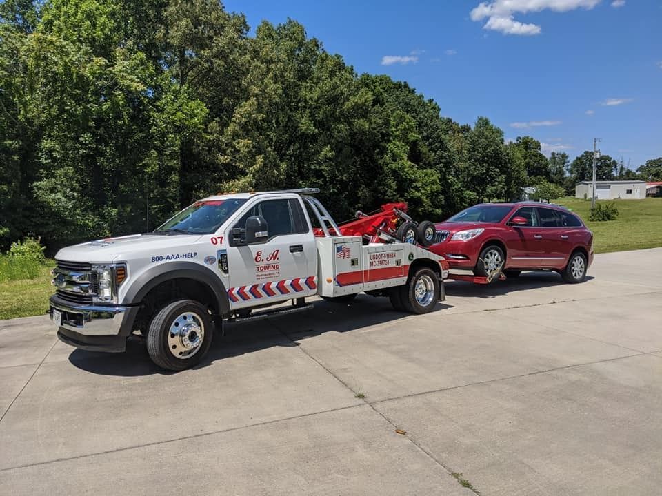 Tow truck towing a red SUV on a sunny day. The truck is white with red accents, parked on asphalt.