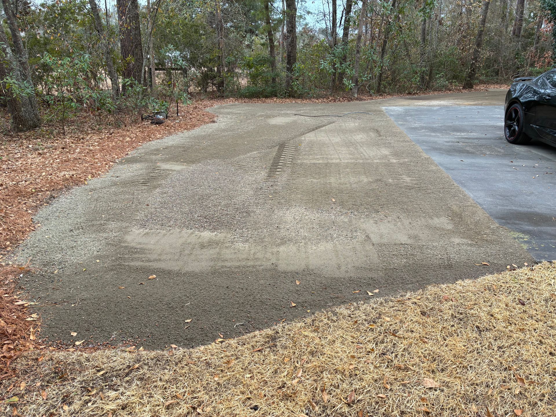 A rectangular patch of packed, grey gravel sits between a wooded area and the edge of a paved driveway.