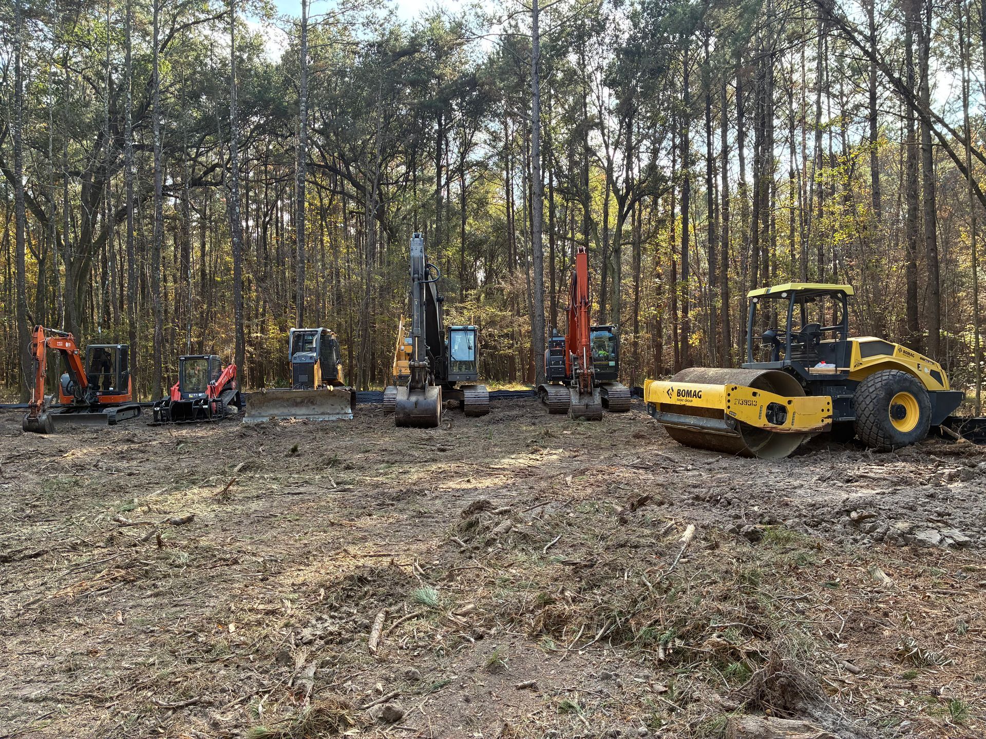 A row of heavy construction machinery, including excavators and a roller, parked in a cleared wooded lot.