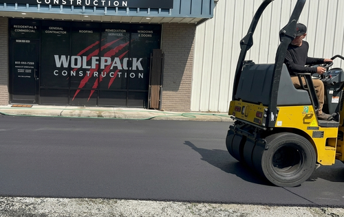 Yellow road roller paving asphalt in front of Wolfpack Construction building
