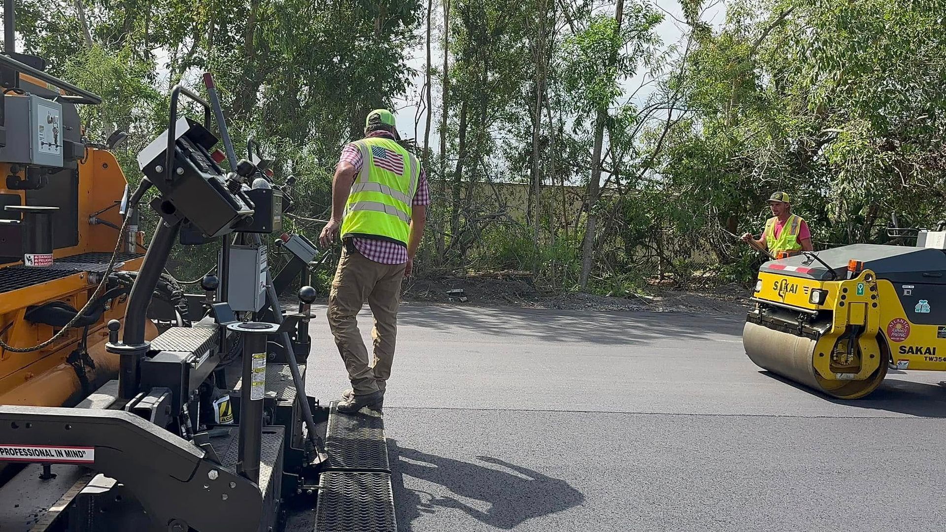 Road work scene with a worker in a reflective vest beside paving machinery and a yellow road roller.
