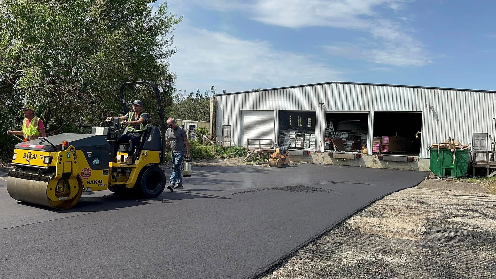 Yellow road roller paving a driveway beside a metal workshop with workers nearby