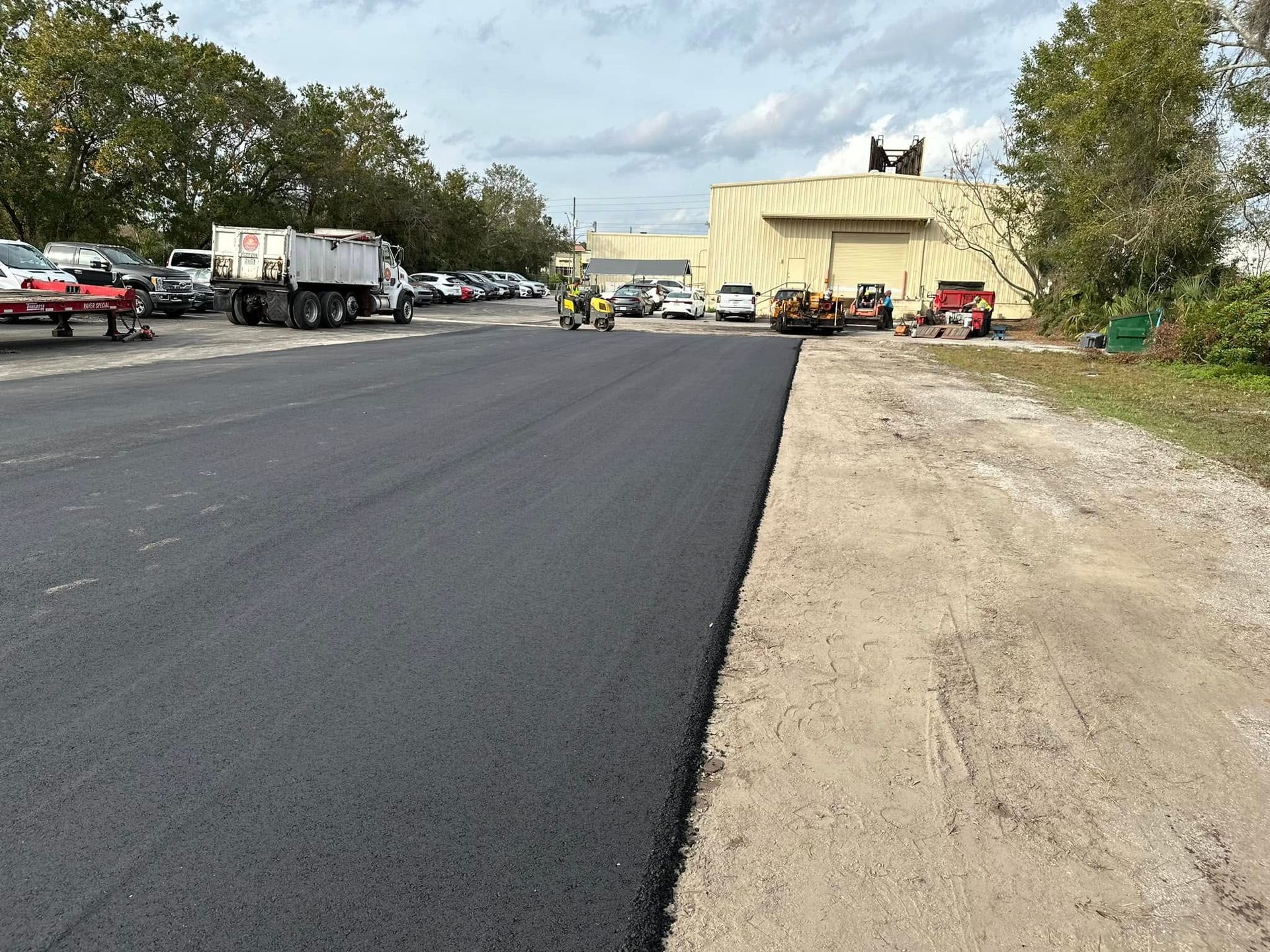 Freshly paved road beside parked trucks and a beige building under cloudy sky
