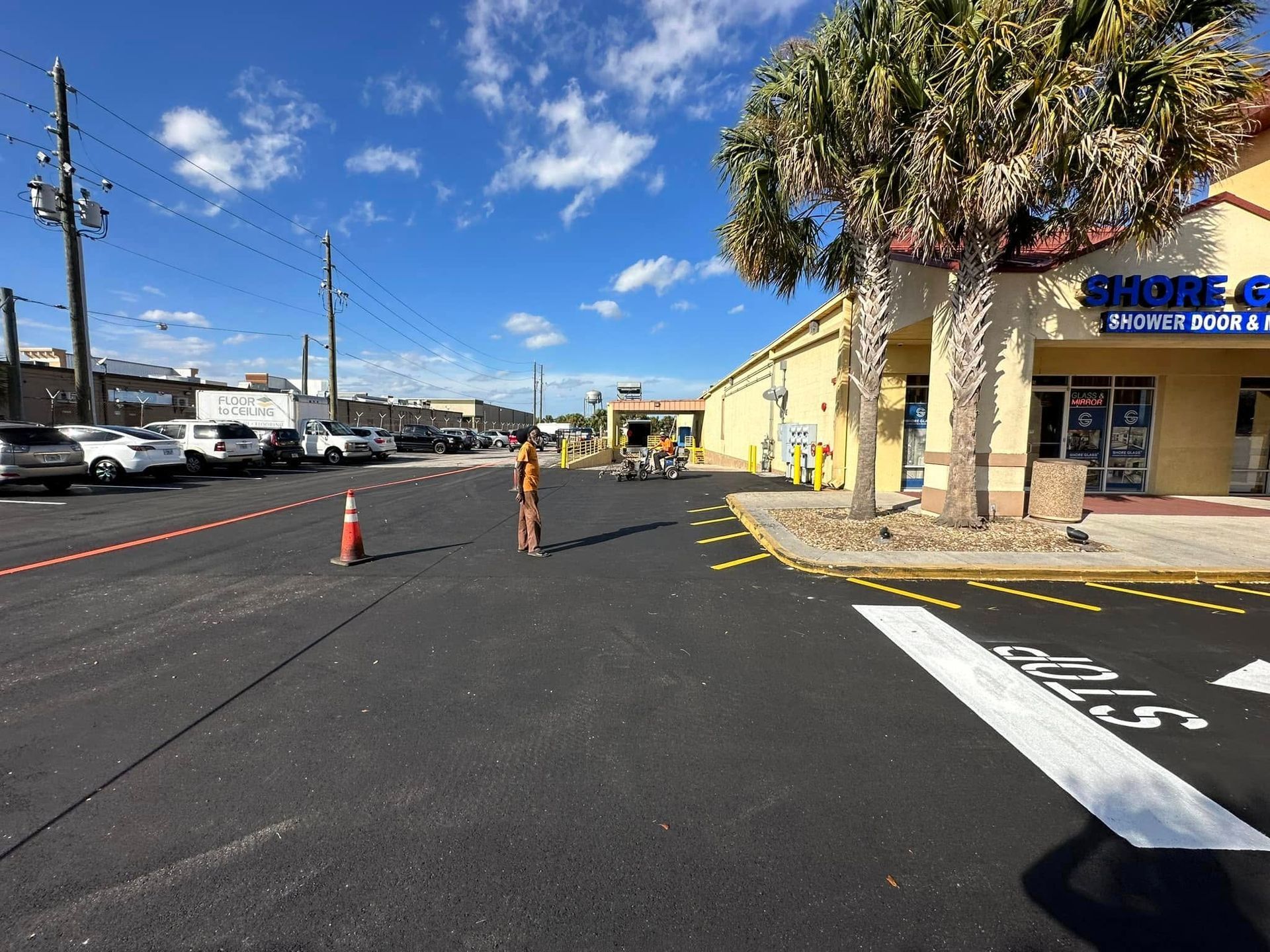 Parking lot beside a yellow storefront with orange cones and a person marking pavement under a blue sky