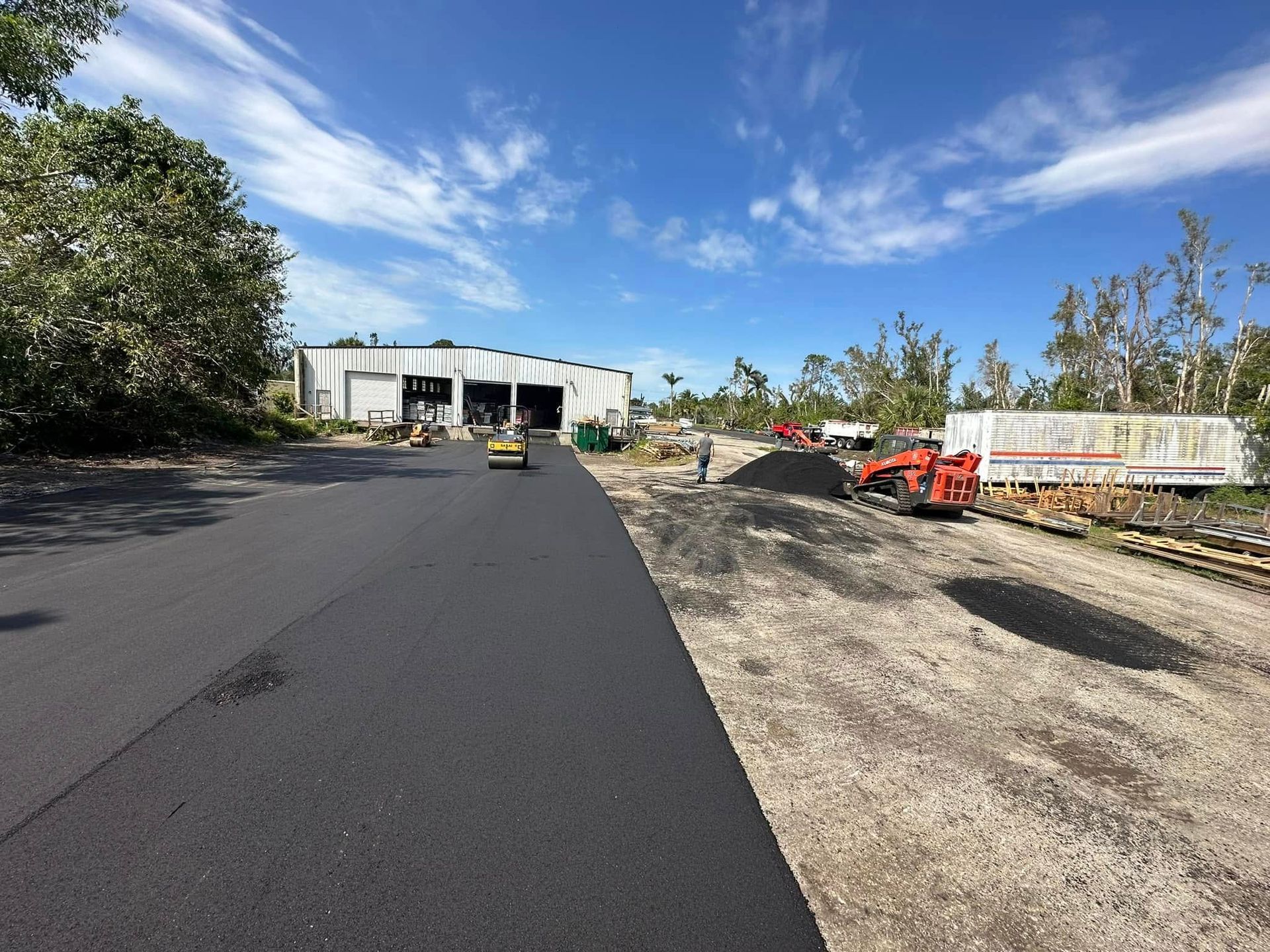 Freshly paved road leading to a warehouse, with construction equipment and gravel roadside under a blue sky