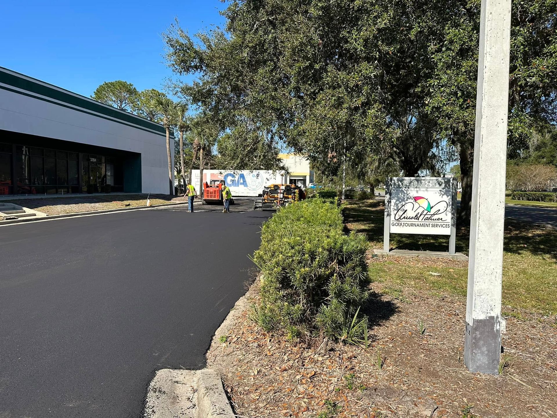 Paved driveway beside a building, with trees, shrubs, and a white utility truck in the distance.