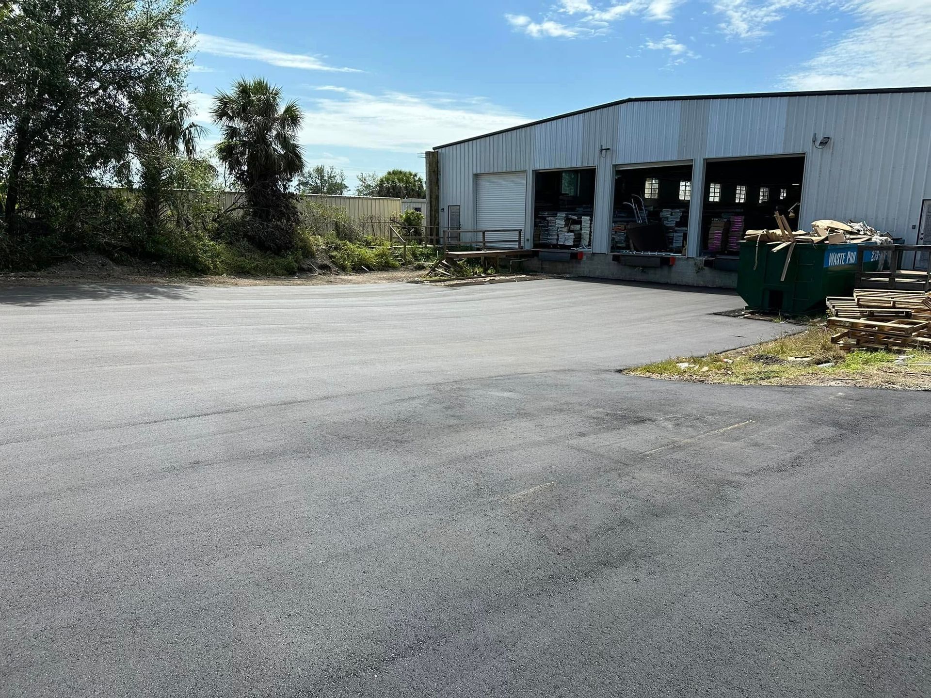 Empty asphalt lot beside a white warehouse with open garage bays under a blue sky