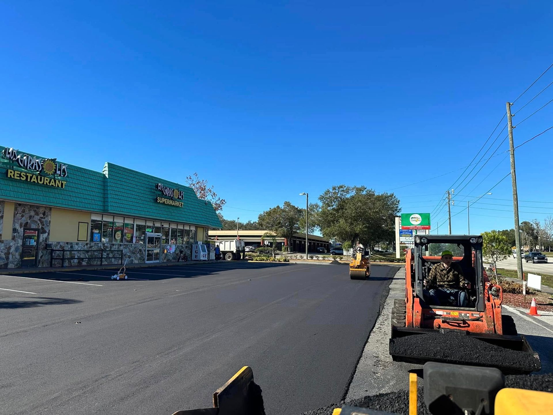 Freshly paved parking lot beside a teal strip mall under a clear blue sky, with construction equipment on the right.