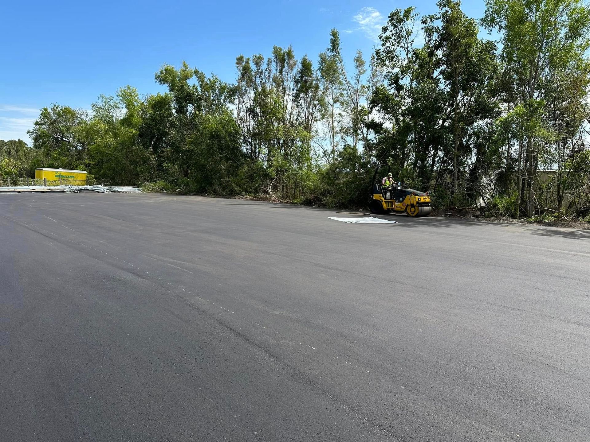Empty paved lot beside trees with yellow construction equipment in the distance