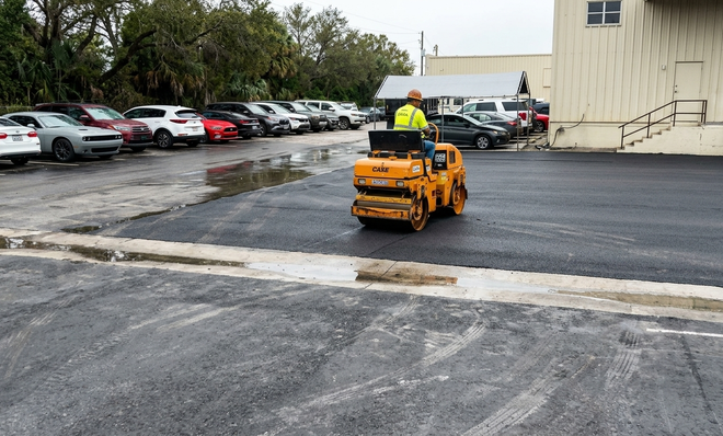 Person operating a small orange roller on a wet parking lot near parked cars and a building