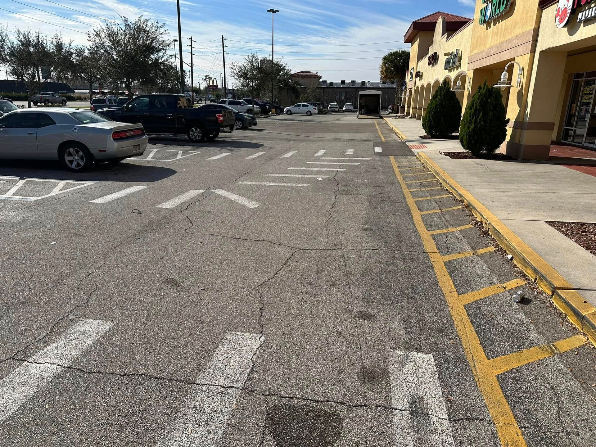 Parking lot beside storefronts with parked cars and a crosswalk under a sunny sky