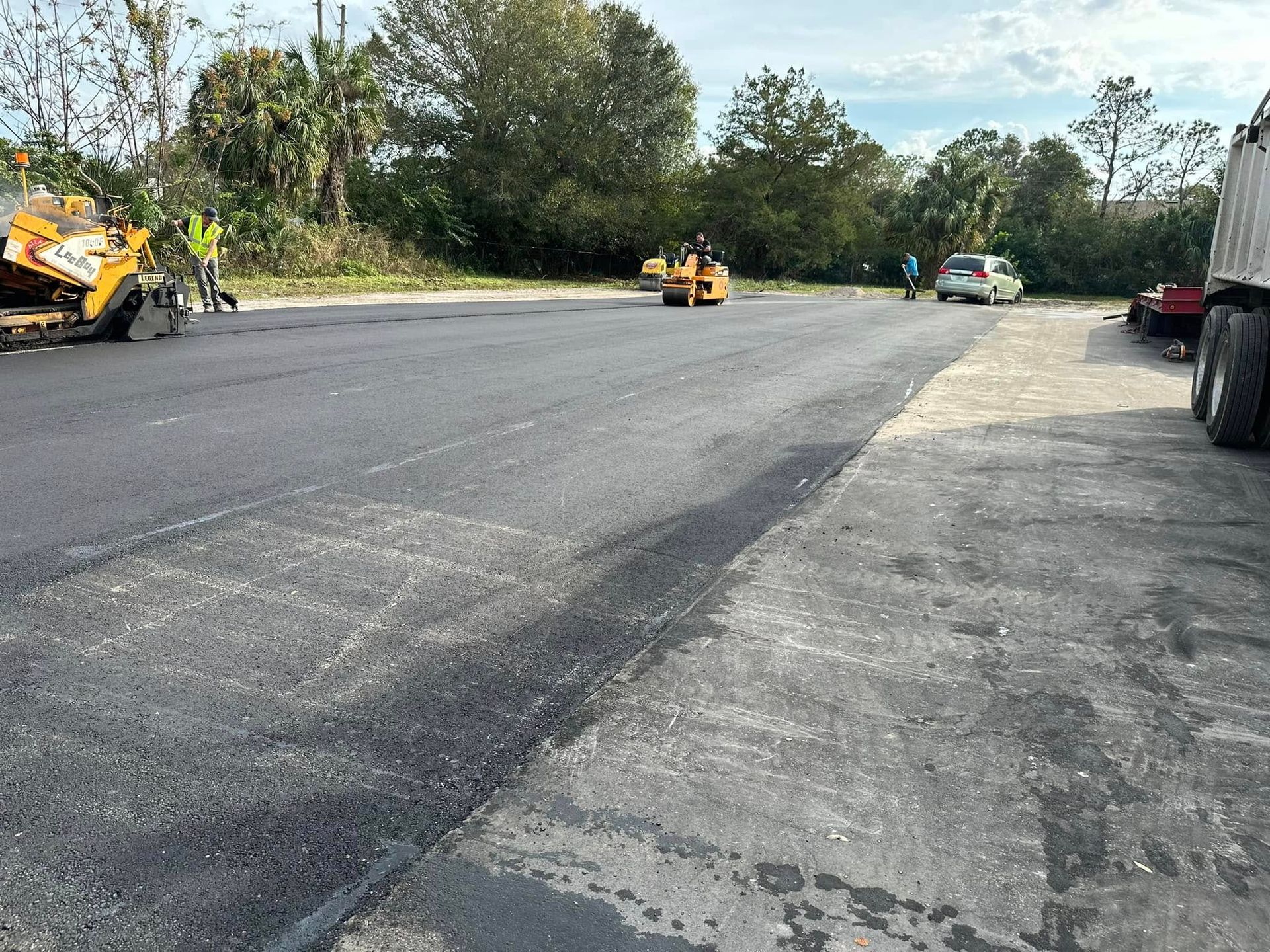 Road paving crew working on a freshly paved road with construction equipment and trees nearby