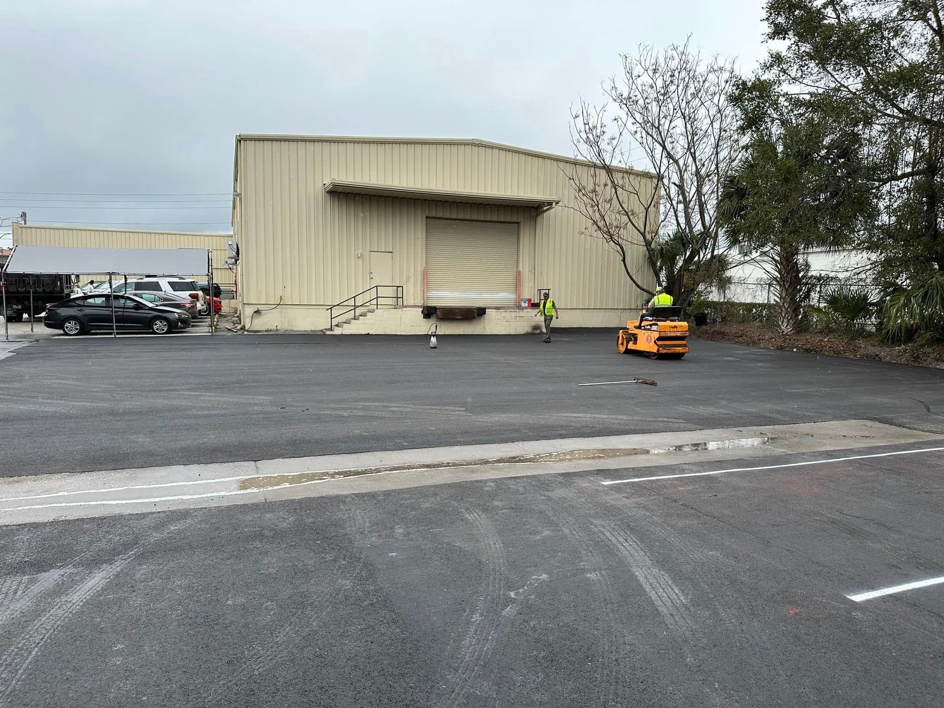 Large empty parking lot beside a beige warehouse, with a small orange vehicle near the loading dock.