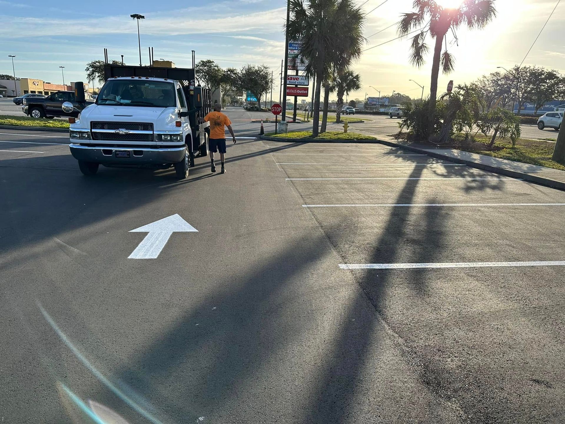 Worker in orange vest pushing a cart beside a white truck in a sunlit parking lot road.