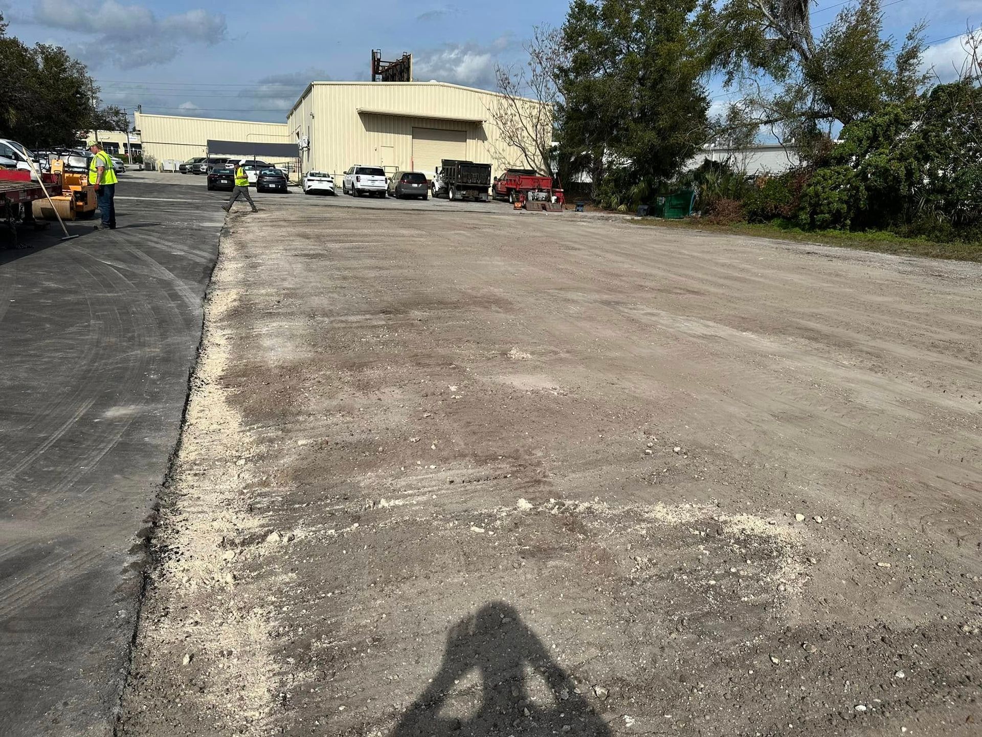 Large empty gravel lot beside a cream building, with parked vehicles and trees under a cloudy sky.