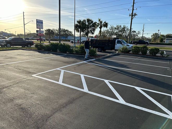 Parking lot with painted white directional lines, median shrubs, and cars by a road at sunset