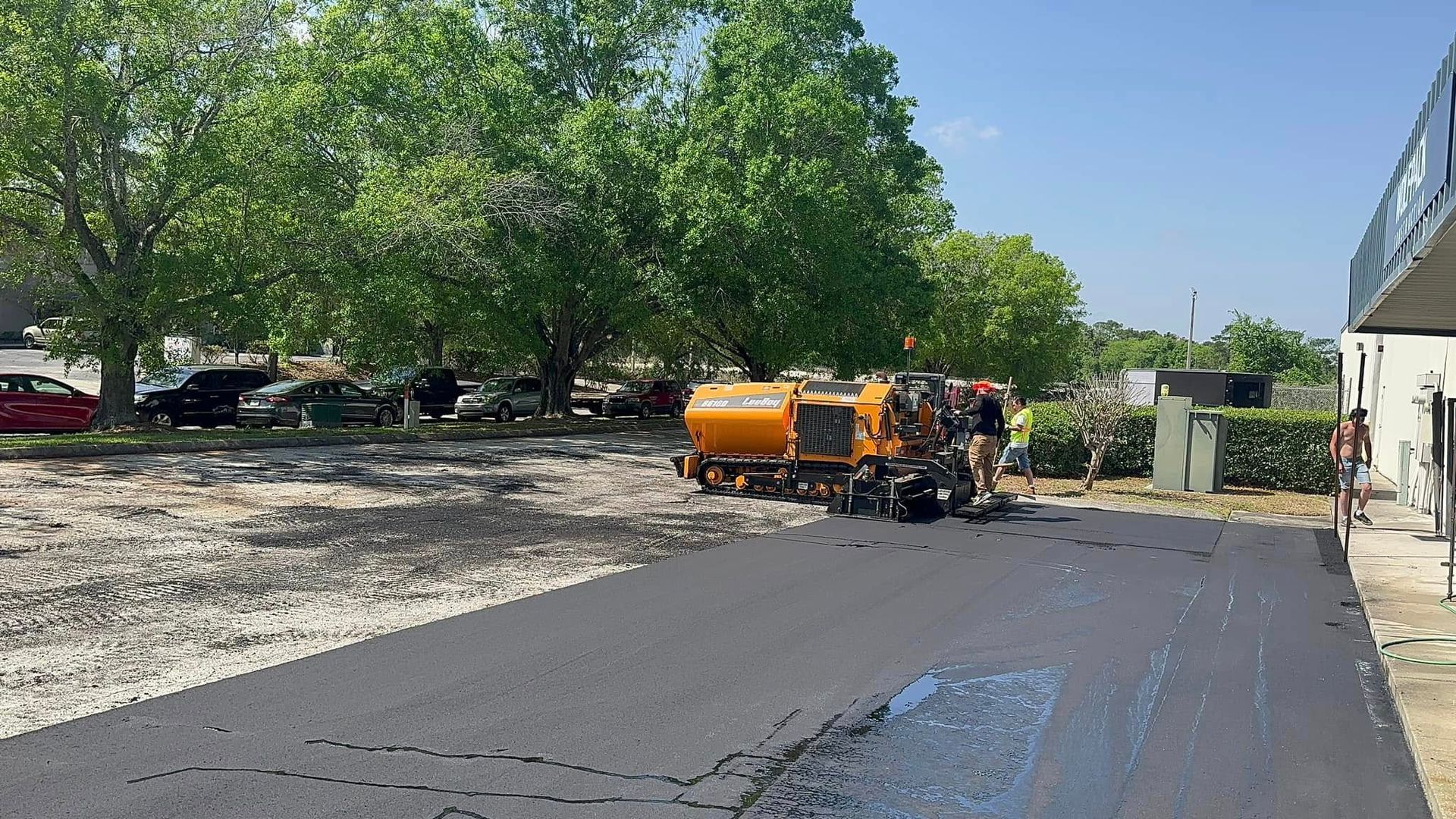 Road paving crew spraying asphalt on a driveway beside a building, with an orange tanker and trees nearby.