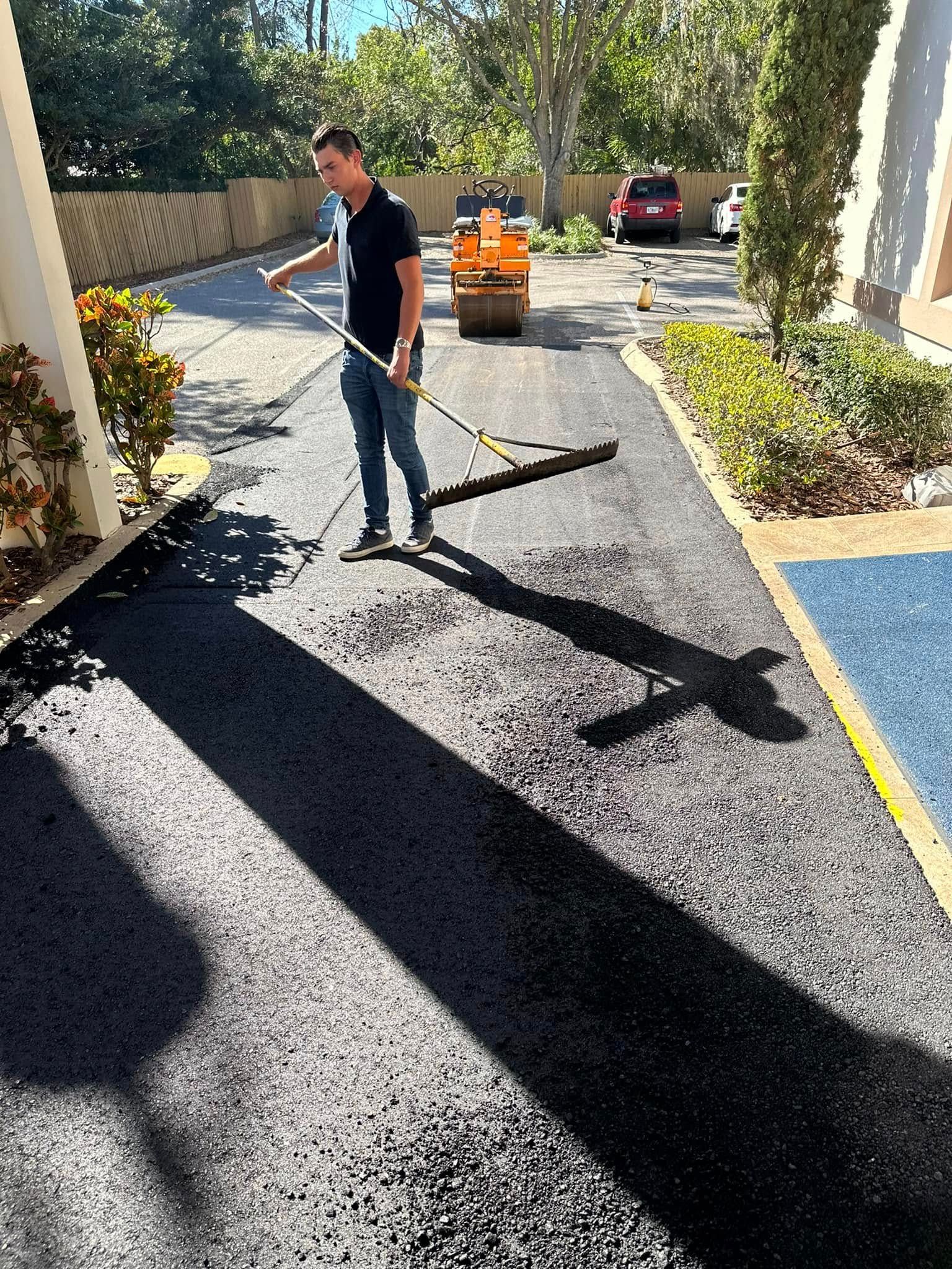Worker raking fresh black asphalt on a driveway, with a roller and car in the background.
