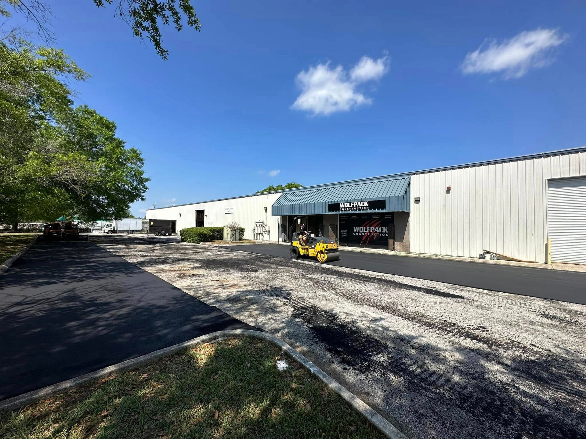 White industrial building with a blue awning, gravel driveway, and trees under a clear blue sky
