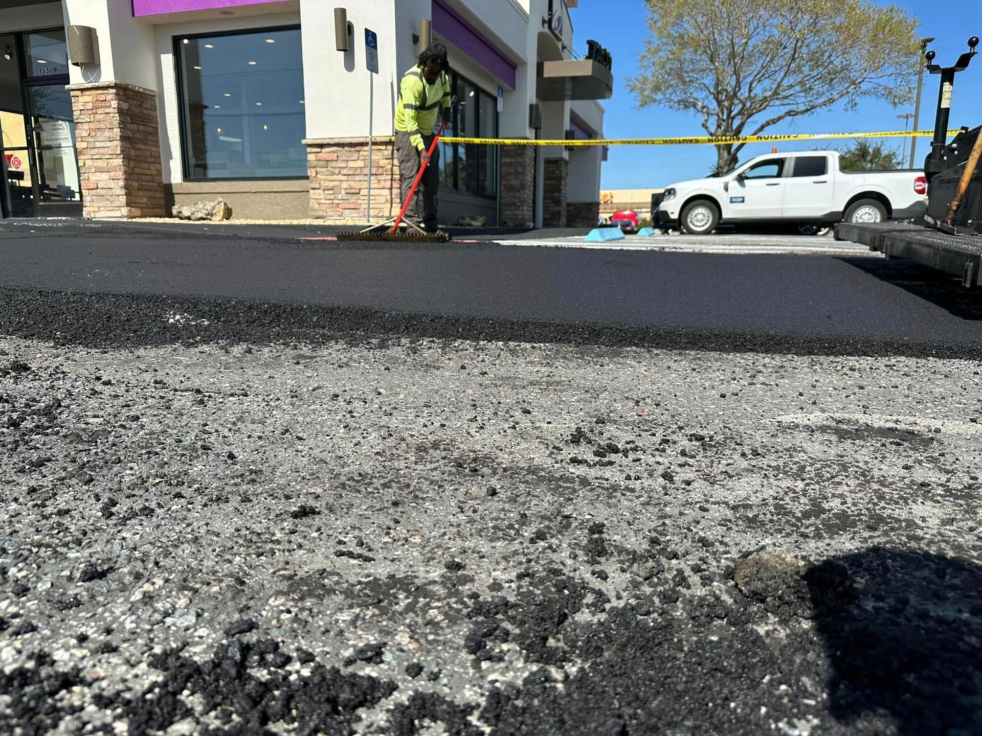 Road resurfacing with workers near a building and parked trucks on a sunny street